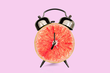 Grapefruit slice on alarm clock, pink pastel background. fruit and vitamins diet at breakfast nutrition concept