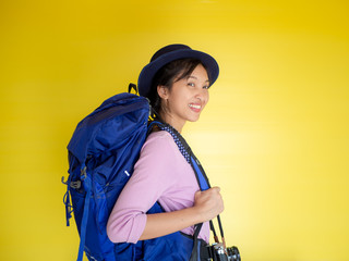 Portrait of a smiling young woman in hat standing with photo camera and looking away at copy space isolated over yellow background,backpack.