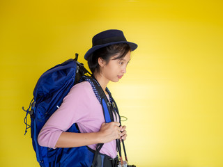 Portrait of a smiling young woman in hat standing with photo camera and looking away at copy space isolated over yellow background,backpack.