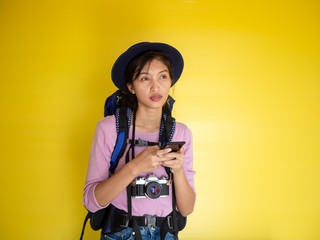 Portrait of a smiling young woman in hat standing with photo camera and looking away at copy space isolated over yellow background,backpack.