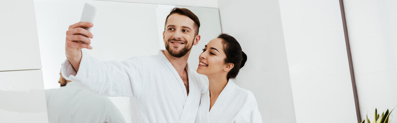 panoramic shot of cheerful man taking selfie while standing with girlfriend in bathroom