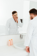 selective focus of upset bearded man looking at mirror in bathroom