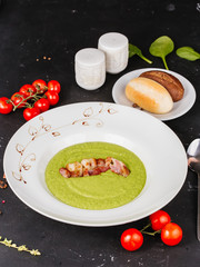 Green pea and bacon puree soup on a table decorated with tomatoes and greens with a bread bun on a black background