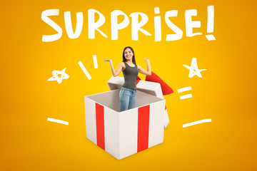 Young smiling woman in sleeveless top and blue jeans appearing from big white gift box on yellow...