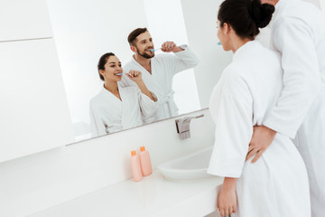 cheerful couple looking at mirror and brushing teeth while standing in bathrobes