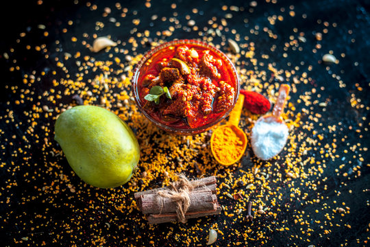 Popular Indian & Asian pickle of mango in a glass bowl i.e. Aam ka achar in a glass bowl or keri ka achar with its entire raw ingredients including spices and mango on black colored shiny surface.