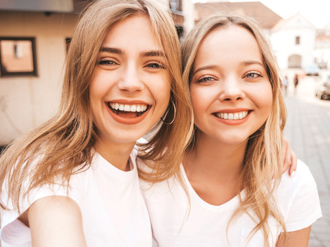 Two young smiling hipster blond women in summer white t-shirt clothes. Girls taking selfie self portrait photos on smartphone.Models posing on street background.Female showing positive face emotions