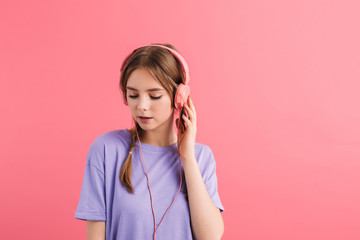 Young attractive girl with two braids in lilac t shirt listening music in headphones dreamily looking aside over pink background isolated