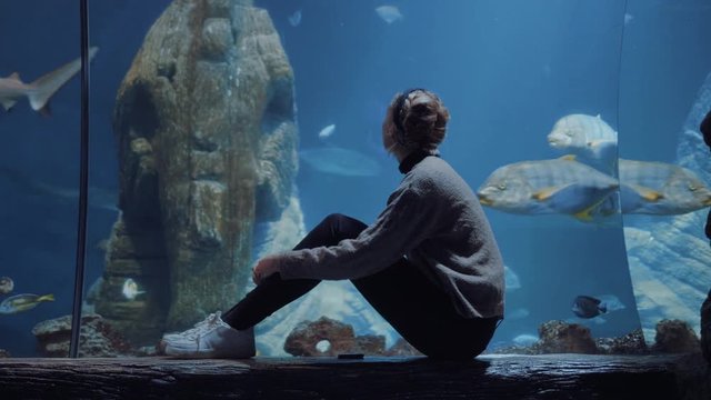 A young blonde woman sits in front of a sea life museum glass while a bunch of different fish swim past her.