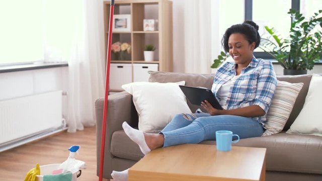 household and technology concept - happy african american woman or housewife with tablet pc computer drinking coffee and resting after home cleaning