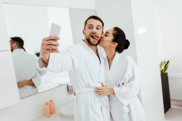 girlfriend kissing cheek of cheerful man showing tongue while taking selfie in bathroom