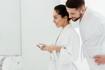 cheerful bearded man looking at happy woman holding toothbrush and toothpaste