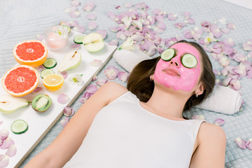 Obraz na płótnie Canvas Beautiful young woman with pink alginate facial mask on her face and slices of fresh cucumber on her lying on the bed in spa. Skin care and treatment, spa, natural beauty and cosmetology concept