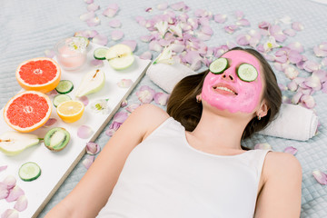 Obraz na płótnie Canvas Beautiful young woman with pink alginate facial mask on her face and slices of fresh cucumber on her lying on the bed in spa. Skin care and treatment, spa, natural beauty and cosmetology concept