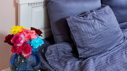 Bedside table with vase of flowers roses and bedroom fresh air. Bed with blue linens, blanket, pillows for comfortable sleep time. Flower interior design and aroma florist concept. Selective focus.