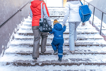 A young family is a little boy and parents return home on transition in the winter in the city. Against the backdrop of snow and stairs. View from the rear, warm winter clothes and shoulder bags.