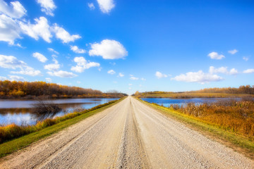 A gravel backroad dividing ponds of water sided with autumn colored trees in a prairie daytime landscape
