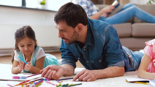 family, fatherhood and leisure concept - father spending time with his little daughters drawing in sketchbooks by crayons and lying on floor at home
