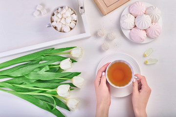 Woman's hands holding cup of tea on the white table with sweets, book and white tulips. Romantic mood. March 8 celebration