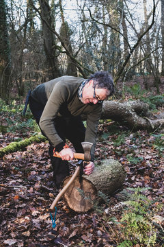 Man standing in forest, using axe and hatchet to split tree trunk.