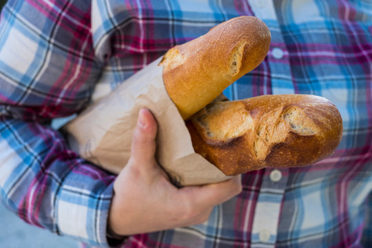 Close up of person holding two freshly baked French baguettes in brown paper bag.