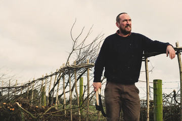 Smiling bearded man holding axe and bill hook standing next to a newly built traditional hedge.