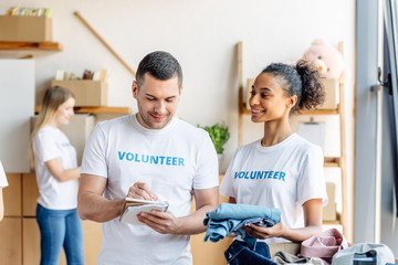 selective focus of smiling multicultural volunteers writing in notebook and unpacking box with clothes