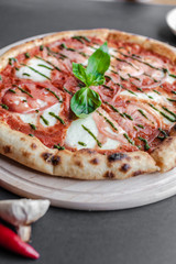 Pizza Margherita with green salsa and leaf of basyl on wooden plate, hot pepper and fried garlic near. Black background, top view, flat lay, vertical image. Natural light.