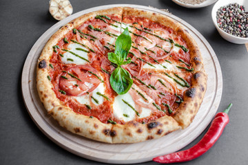 Pizza Margherita with green salsa and leaf of basyl on wooden plate, garlic, red pepper and bowl with spices near. Black background, top view, flat lay, vertical image. Natural light.