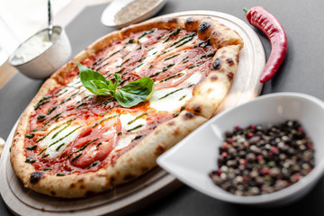 Pizza Margherita on wooden plate with red pepper and white bowl with spices near, everything on the table near the window. Natural light. Shallow focus. Horizontal image.