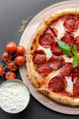 Pepperoni pizza with leaf of basyl on the wooden plate, plate with salami, cherry tomato branch and cumin near. Top view, flat lay, black background. Copy space, vertical image. Natural light. 