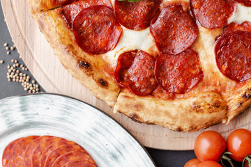 Close up Pepperoni pizza on the wooden plate, plate with salami, cherry tomato branch, cumin near. Top view, flat lay, black background. Horizontal image. Natural light. 