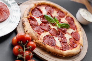 Close up Pepperoni pizza with basyl leaf in the middle on the wooden plate, plate with salami slices, cherry tomato branch, bowl with flour near. Black background. Horizontal image. Natural light. 