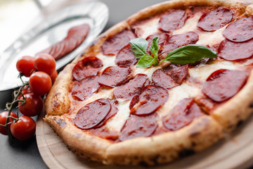 Close up Pepperoni pizza with basyl leaf in the middle on the wooden plate, plate with salami slices, cherry tomato branch near. Black background. Horizontal image. Natural light. 