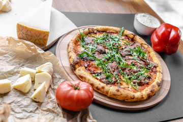 Pizza with roast beef, arugula on the wooden plate. Tomato, bell pepper and pieses of cheese near on the table. Horizontal. Natural light. Black background. Shallow focus.