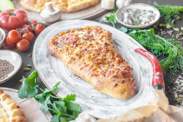 Georgian khachapuri pie with cheese and meat on the plate, red pepper, tomatoes, mushrooms, herbs, onion and bowls with spices nearby. Black background. Horizontal image. 