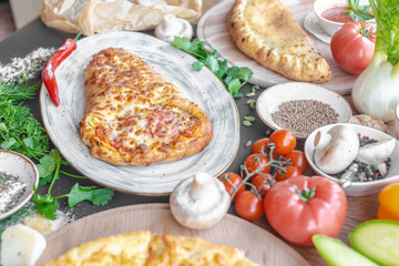 Georgian khachapuri pie with cheese and meat on the plate, red pepper, tomatoes, mushrooms, herbs, onion and bowls with spices nearby. Black background. Horizontal image. 