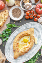 Georgian national pie khachapuri with egg, cheese and butter on the plate, parsley, dill, bowl with caraway, vegetables, and another pie near. Vertical image. Top view. Black background