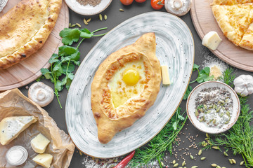 Georgian national pie khachapuri with egg, cheese and butter on the plate, parsley, dill, bowl with spices, cheese pieses, tomatoes, muchrooms and another pies near. Horizontal image. Black background