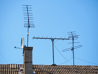 Satellite and old television antennas are installed on the sloping slate roof of the house. Antennas are attached with cables. Rooftop chimney. Against the backdrop of a clean blue sky.