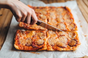 Female hand cut homemade square pizza with smoked sausage and cheese. White papyrus, wooden background, close up.
