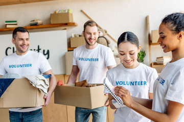 four young multicultural volunteers in white t-shirts with volunteer inscriptions working in charity center