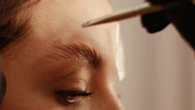 Close up of female cosmetologist covers the eyebrows of a beautiful patient with a special remedy. Part of eyebrow lamination procedure