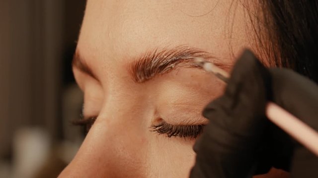 Close-up of cosmetologist hand applys paint with brush on the eyebrows of a beautiful patient. Part of eyebrow correction procedure. Professional care for face in beauty saloon