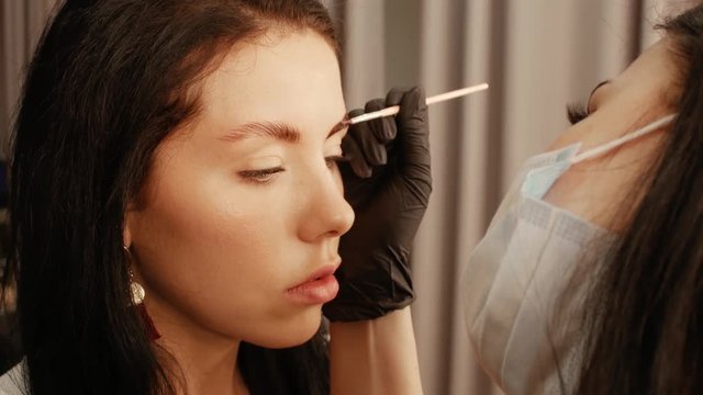 Woman cosmetologist apply paint with brush on the eyebrows of a beautiful patient. Part of eyebrow correction procedure close-up. Professional care for face in beauty saloon