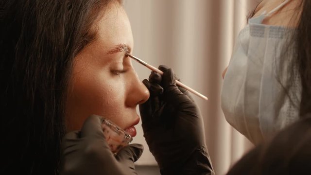 Female cosmetologist apply paint with brush on the eyebrows of a beautiful patient. Part of eyebrow correction procedure close-up. Professional care for face in beauty saloon