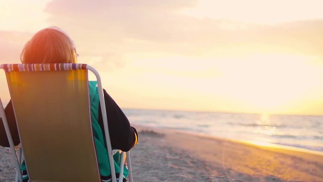 Empty sandy beach at sunrise/sunset. The girl sits on a sun lounger, looks at the sea and listens to music on headphones. Fixed Camera
