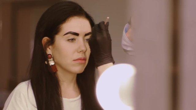 Cosmetologist apply paint with brush on the eyebrows of a beautiful patient. Part of eyebrow correction procedure. Professional care for face in beauty saloon