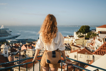 Fototapeta na wymiar Blonde woman standing on the balcony and looking at coast view of the southern european city with sea during the sunset, wearing hat, cork bag, safari shorts and white shirt