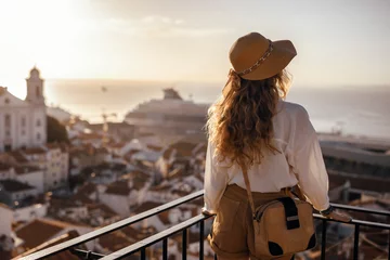 Papier Peint photo Lavable Madrid Blonde woman standing on the balcony and looking at coast view of the southern european city with sea during the sunset, wearing hat, cork bag, safari shorts and white shirt